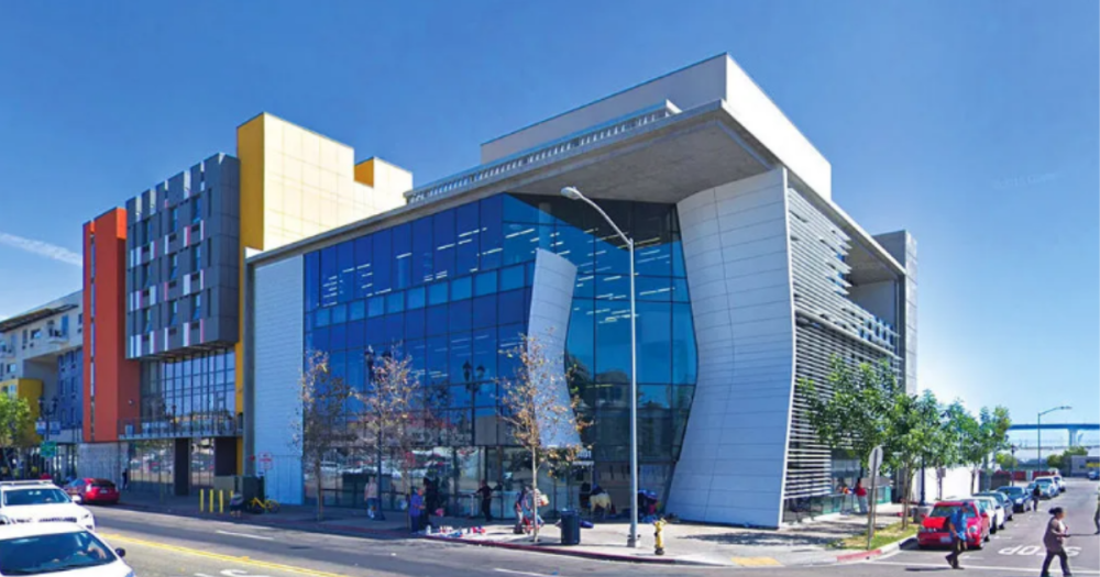 A glass building located at the corner of a street surrounded by people walking on the sidewalk and parked cars