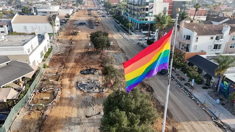 A large rainbow Pride flag flies over a street construction site in Hillcrest, with homes and buildings lining both sides of the road.