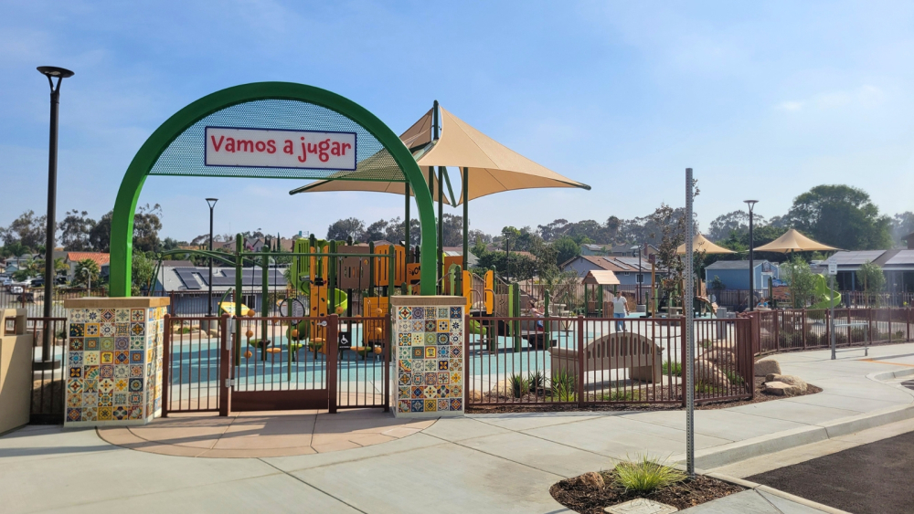 A colorful playground with climbing structures and slides is visible behind a gate with the sign "Vamos a jugar." The playground is surrounded by decorative tile walls and shaded by large canopies. Houses and trees are in the background under a clear blue sky.