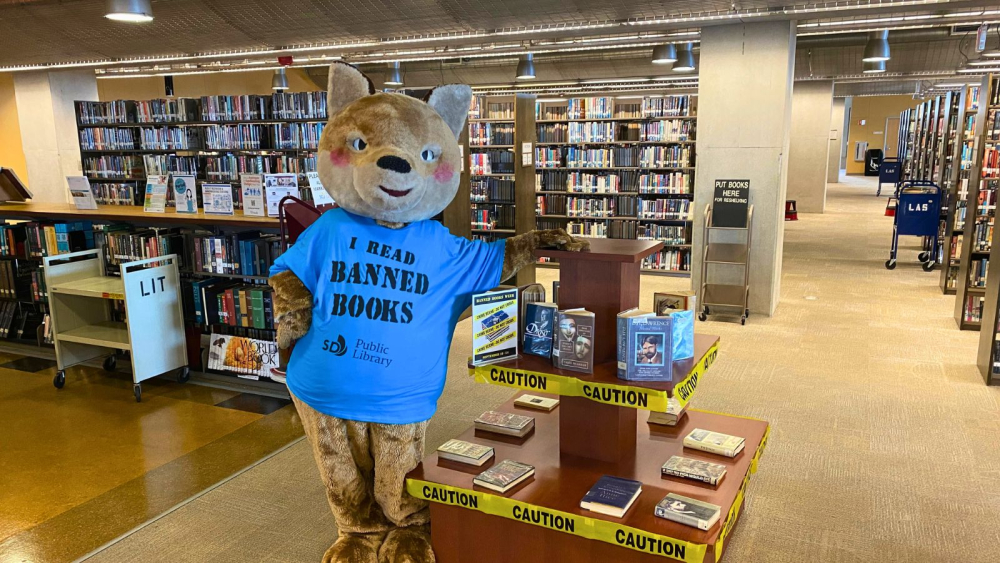 A large bear mascot wearing a blue shirt with the text "I Read Banned Books" stands in a library. The shirt also displays a "Public Library" logo. The mascot is beside a display of books surrounded by yellow caution tape. Bookshelves line the background.