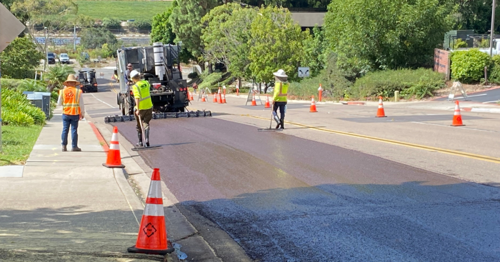 three workers slurry sealing a section of a street, surrounded by trees