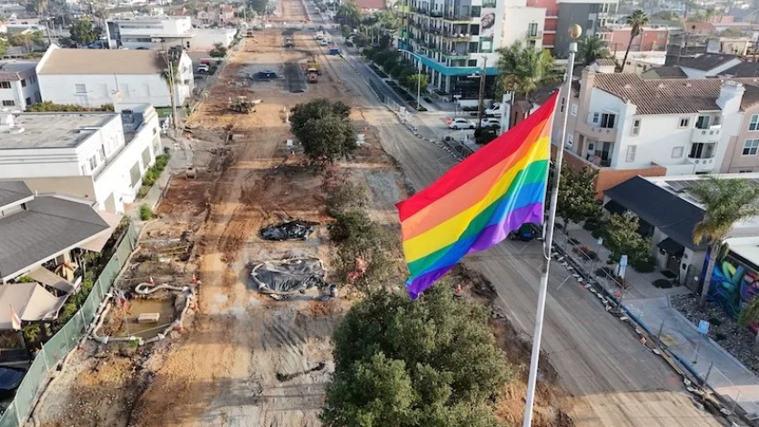 A large rainbow Pride flag flies over a street construction site in Hillcrest, with homes and buildings lining both sides of the road.