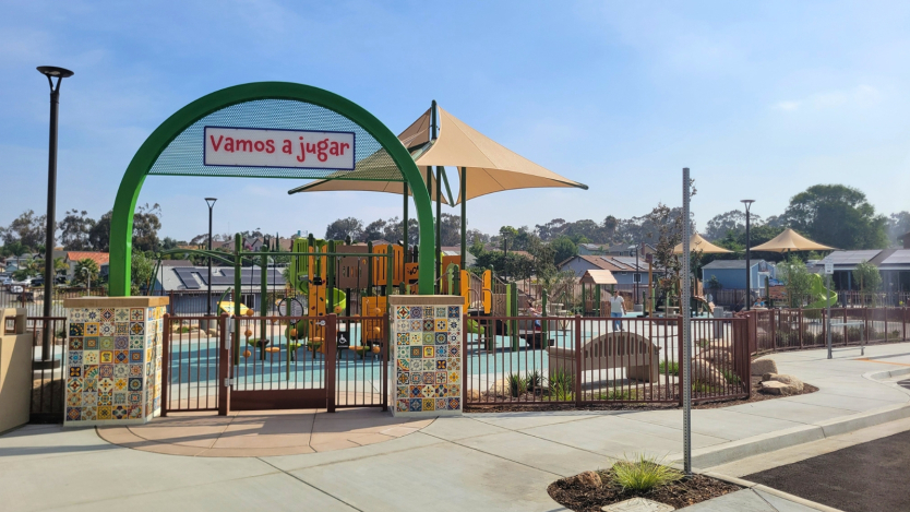 A colorful playground with climbing structures and slides is visible behind a gate with the sign "Vamos a jugar." The playground is surrounded by decorative tile walls and shaded by large canopies. Houses and trees are in the background under a clear blue sky.