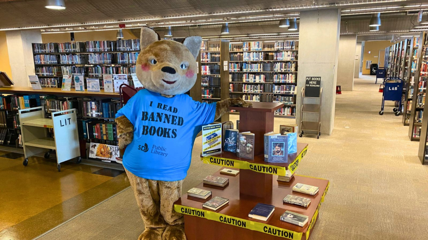 A large bear mascot wearing a blue shirt with the text "I Read Banned Books" stands in a library. The shirt also displays a "Public Library" logo. The mascot is beside a display of books surrounded by yellow caution tape. Bookshelves line the background.
