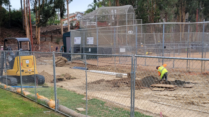 construction worker digging dirt surrounded by a chain fence and baseball field